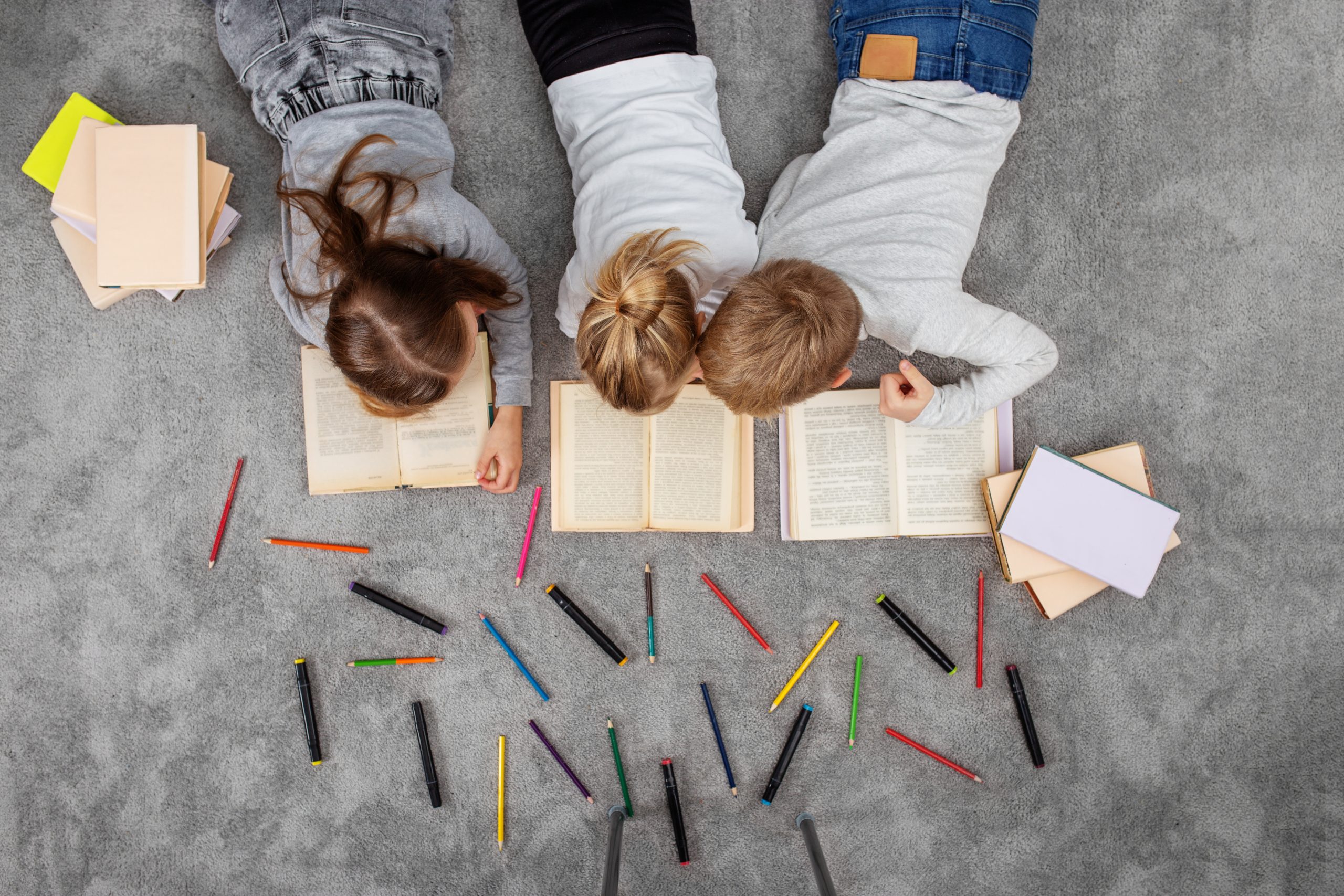 3 kinderen lezen een boek liggend op de grond met kleurpotloden en stiften om hen heen